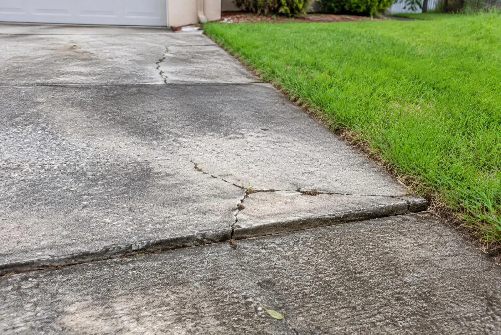 Aging concrete driveway with cracks and an uneven, settled section beside a lawn